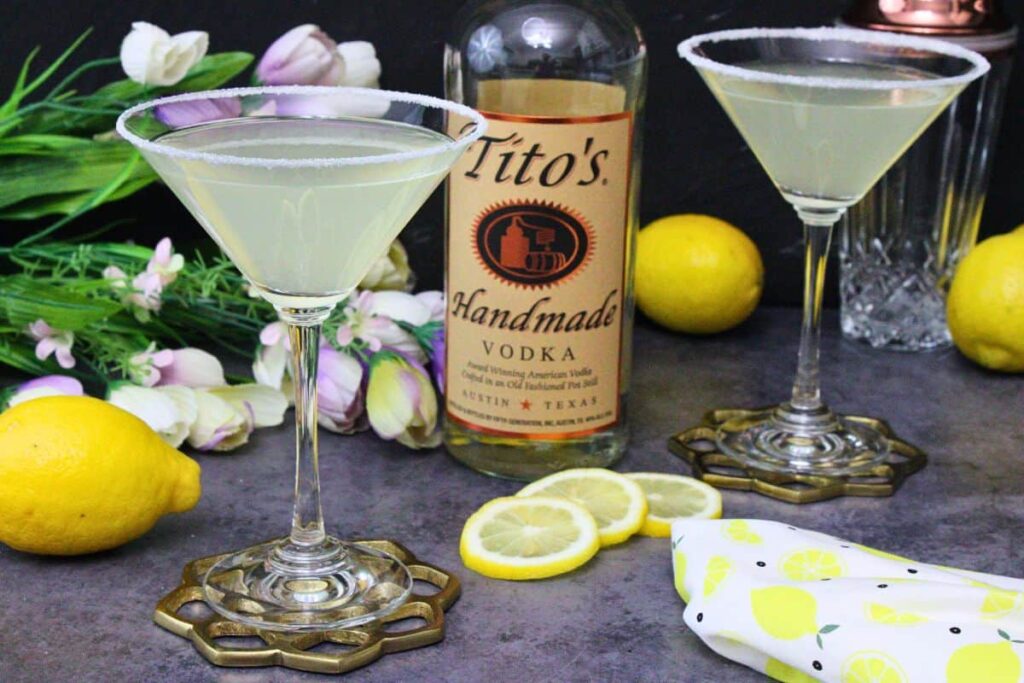 two lemon drop martinis surrounding a bottle of vodka with fresh lemons