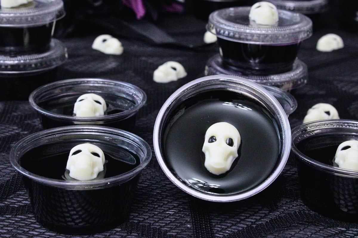 closeup of several black jello shots in plastic cups topped with a white chocolate skull on a black lace web cloth