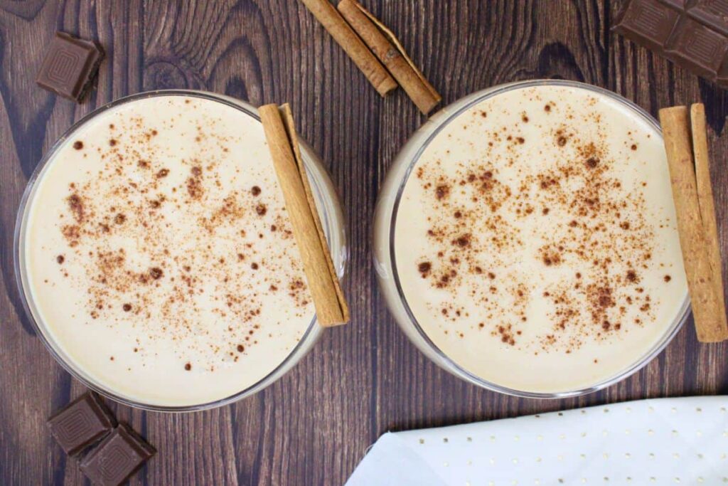 overhead view of two chocolate whiskey flips dusted with cinnamon and cinnamon stick garnish