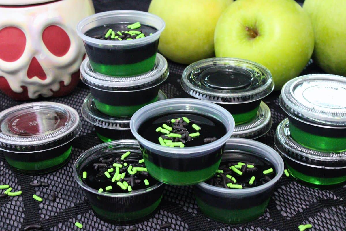 several black and green poison apple jello shots topped with black and green sprinkles with fresh apples in the background