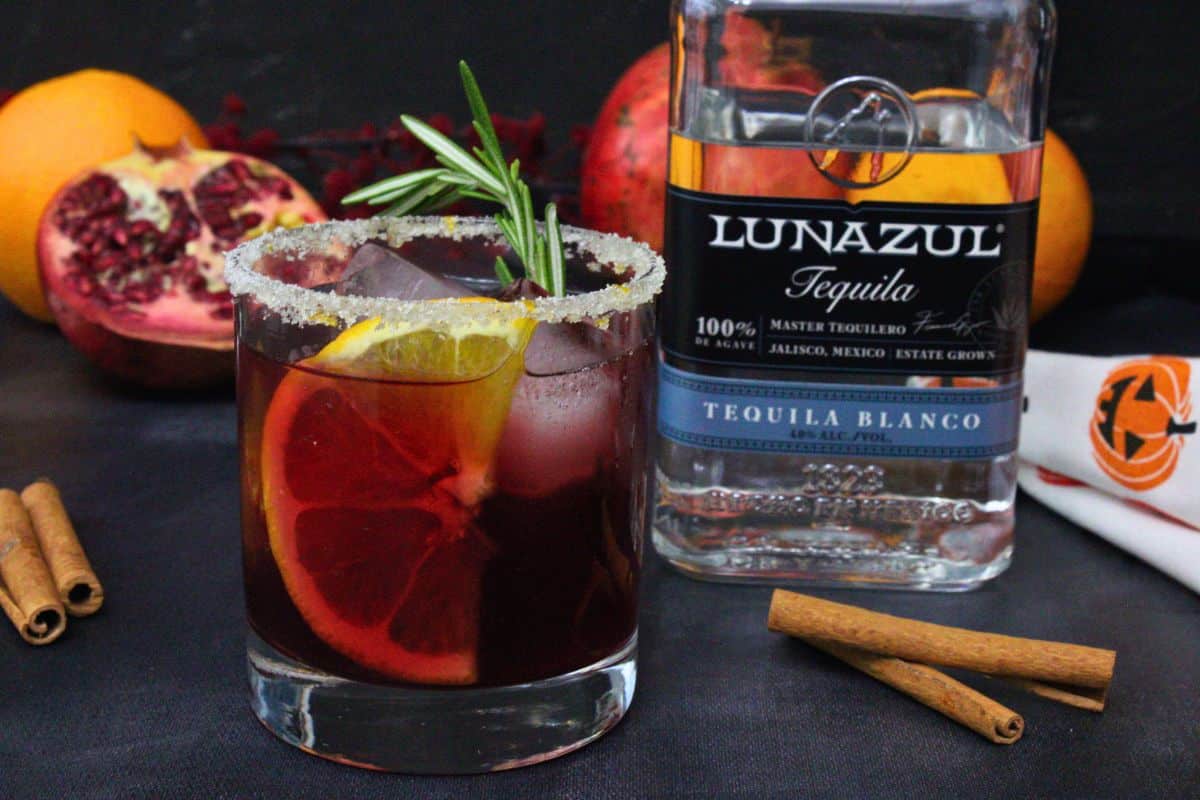 sleepy hollow cocktail with orange slice and rosemary garnish next to tequila bottle against a dark background