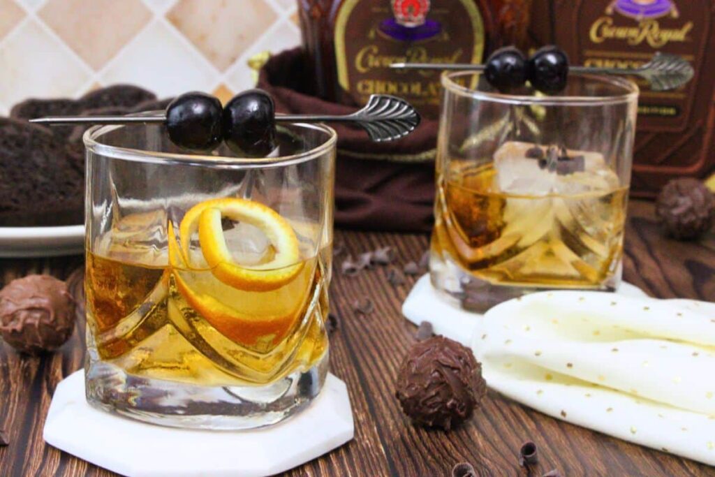 two chocolate old fashioned drinks garnished with orange peel and cocktail cherries in front of chocolate crown royal bottle