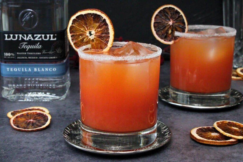 blood orange margarita with blood orange slice and sugar rim on a metal coaster with separate drink and tequila bottle in background