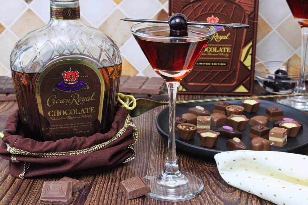 chocolate manhattan cocktail in coupe glass with cherry skewer next to chocolate crown royal bottle and chocolate candies