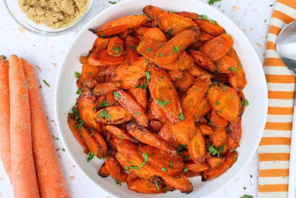 overhead view of air fryer brown sugar carrots in a white bowl next to fresh carrots and a serving spoon
