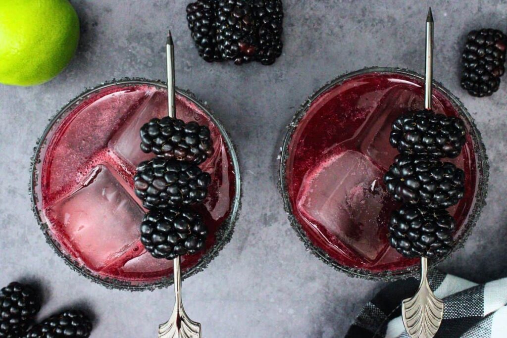 overhead view of two black magic margaritas with blackberry skewer and fresh fruit on black counter