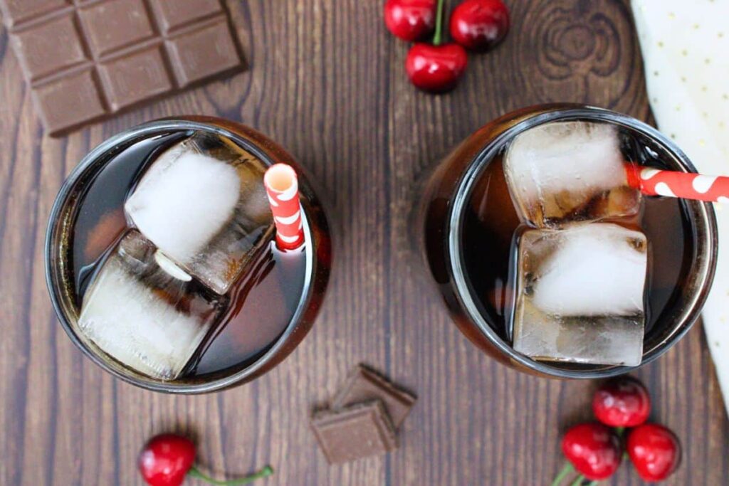overhead view of two crown royal chocolate and cherry coke drinks with ice and paper straws on a wooden counter