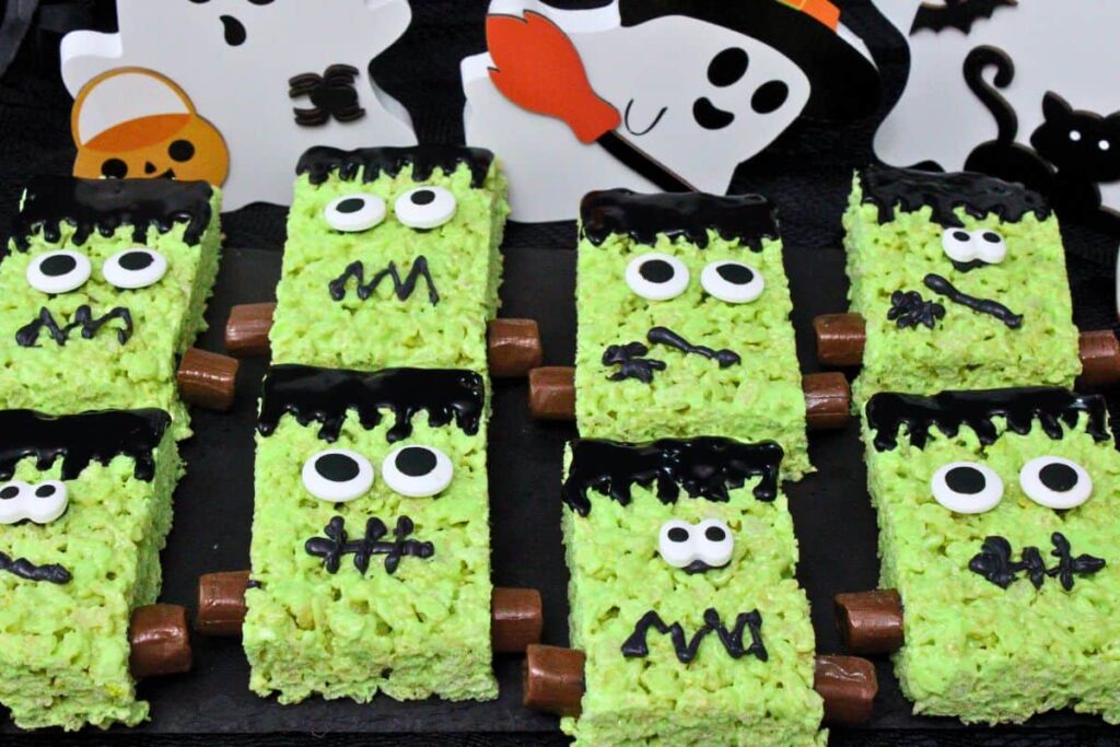 several frankenstein rice krispie treats with candy eyes and tootsie roll bolts on a black slate with ghost decorations