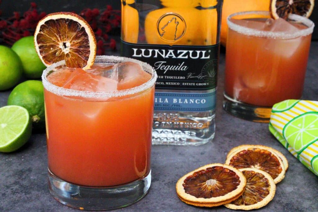 two blood orange margaritas garnished with blood orange slice around tequila bottle and fresh limes