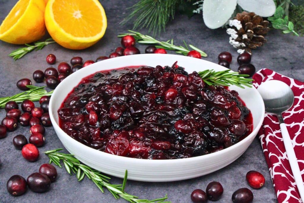 bowl of microwave cranberry sauce with a sprig of rosemary surrounded by fresh cranberries, rosemary, and oranges