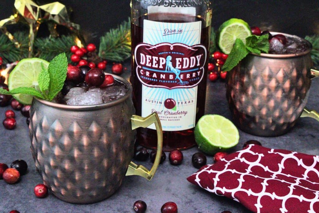 cranberry moscow mule cocktail in a copper mug with mint and lime garnish next to vodka bottle surrounded by fresh cranberries