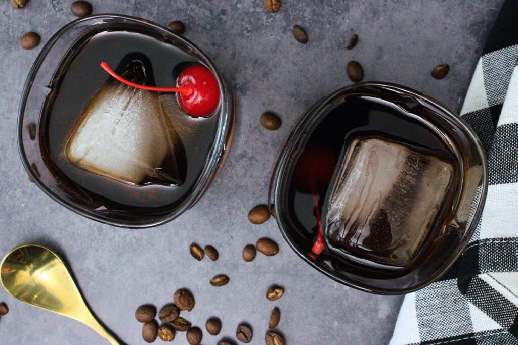overhead view of two black russian cocktails with cherry garnish surrounded by fresh coffee beans
