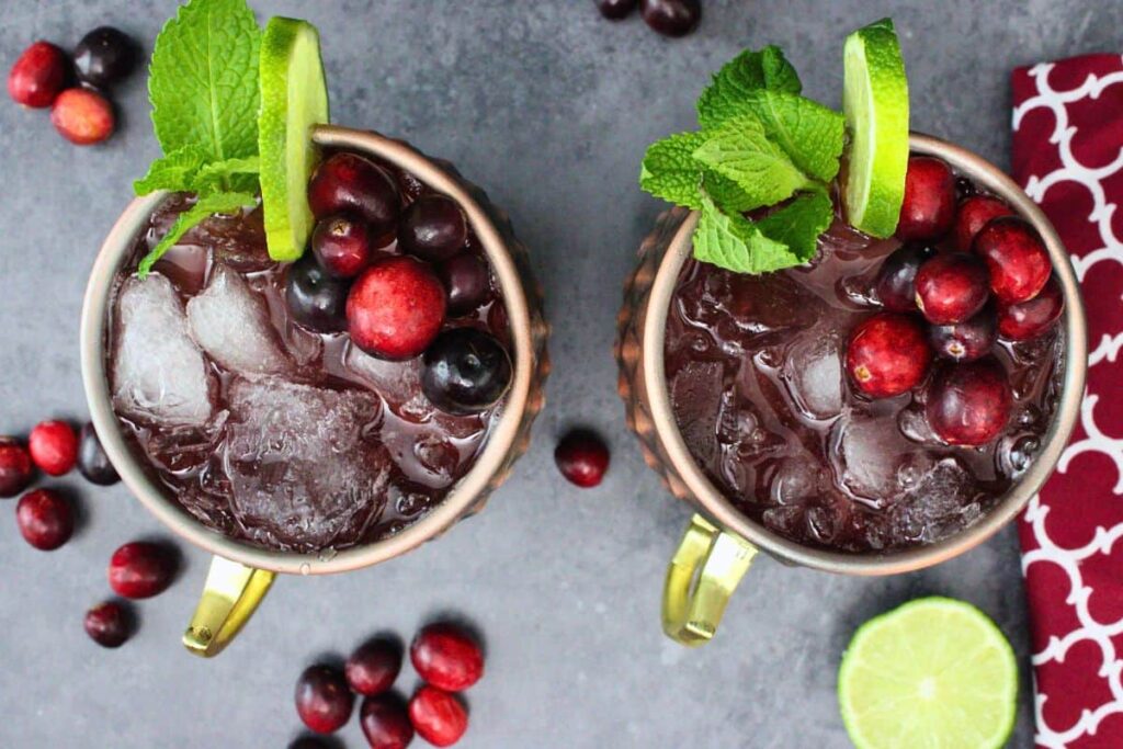 overhead view of two cranberry moscow mule drinks with fresh lime, mint, and cranberry garnish on a dark counter