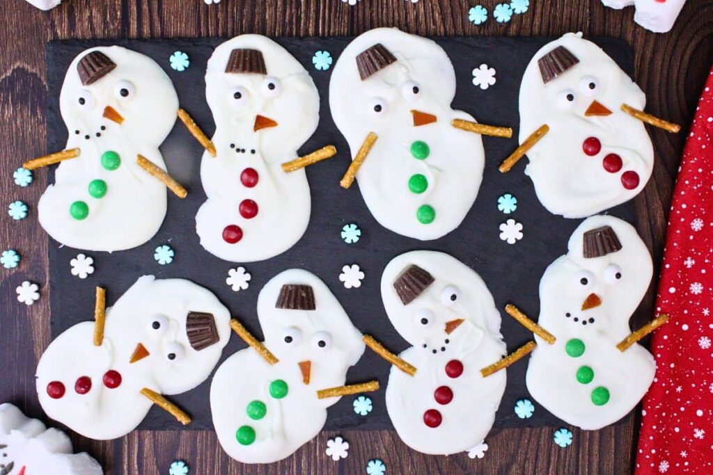overhead view of several pieces of melted snowman bark on a piece of slate with chocolate and candied features plus pretzel arms