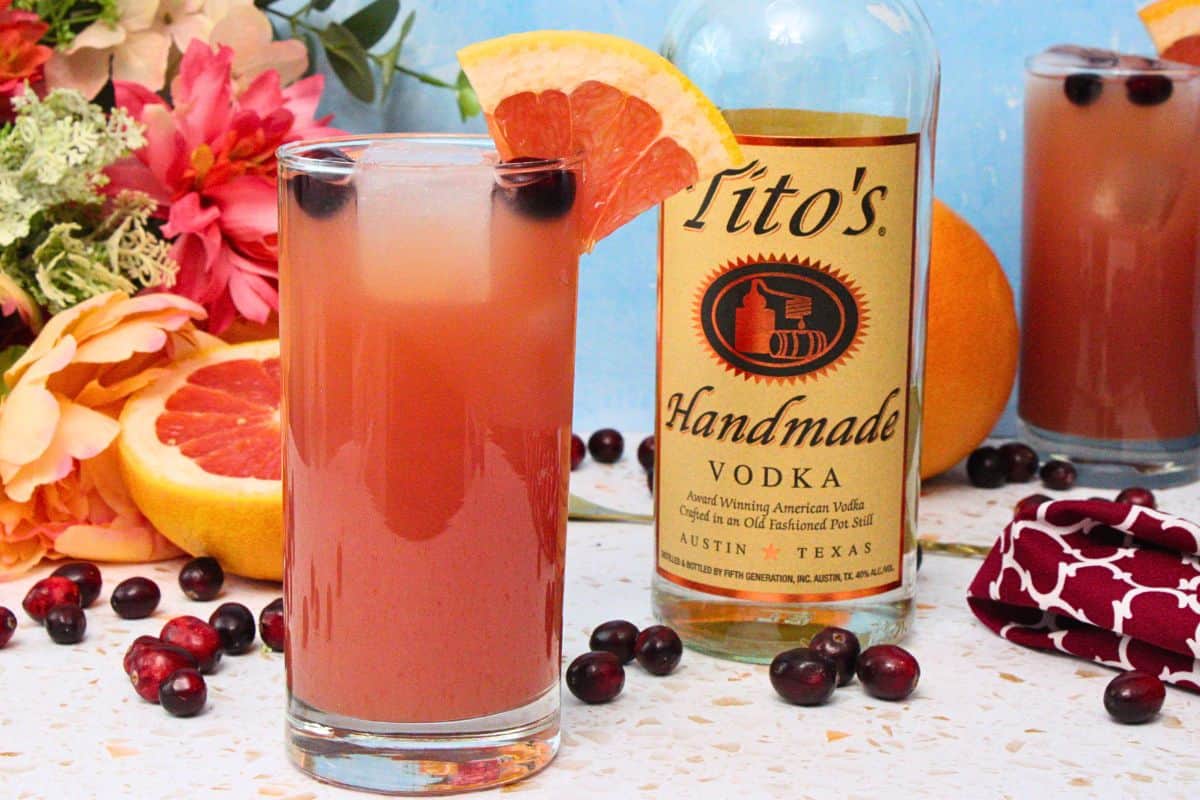 sea breeze cocktail in tall glass with grapefruit wedge garnish next to vodka bottle amidst fresh fruit and flowers