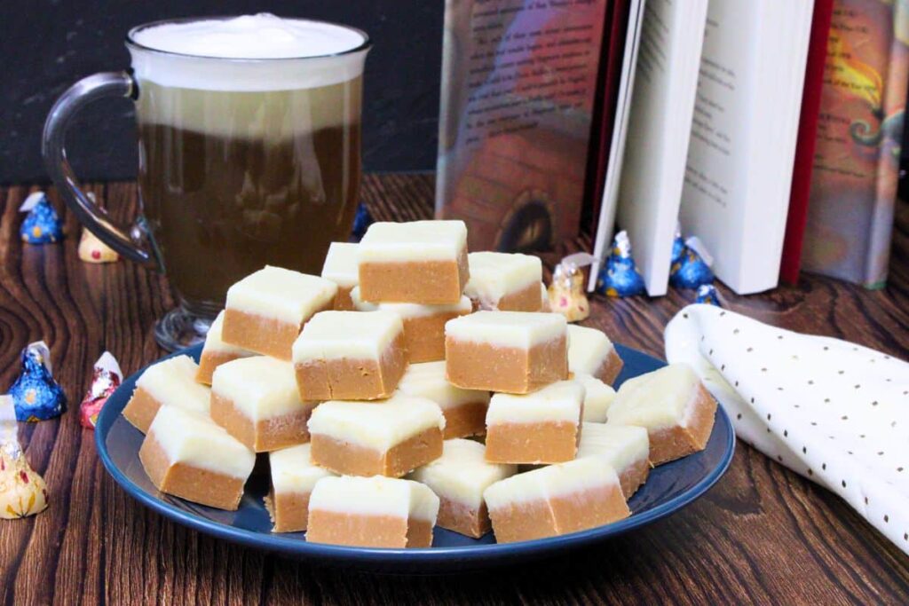 squares of butterbeer fudge on a plate in front of a mug filled with foamy butterbeer and an open harry potter book