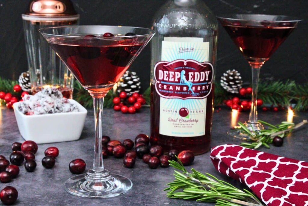 two cranberry martinis with vodka bottle in the center surrounded by fresh cranberries and pine sprigs