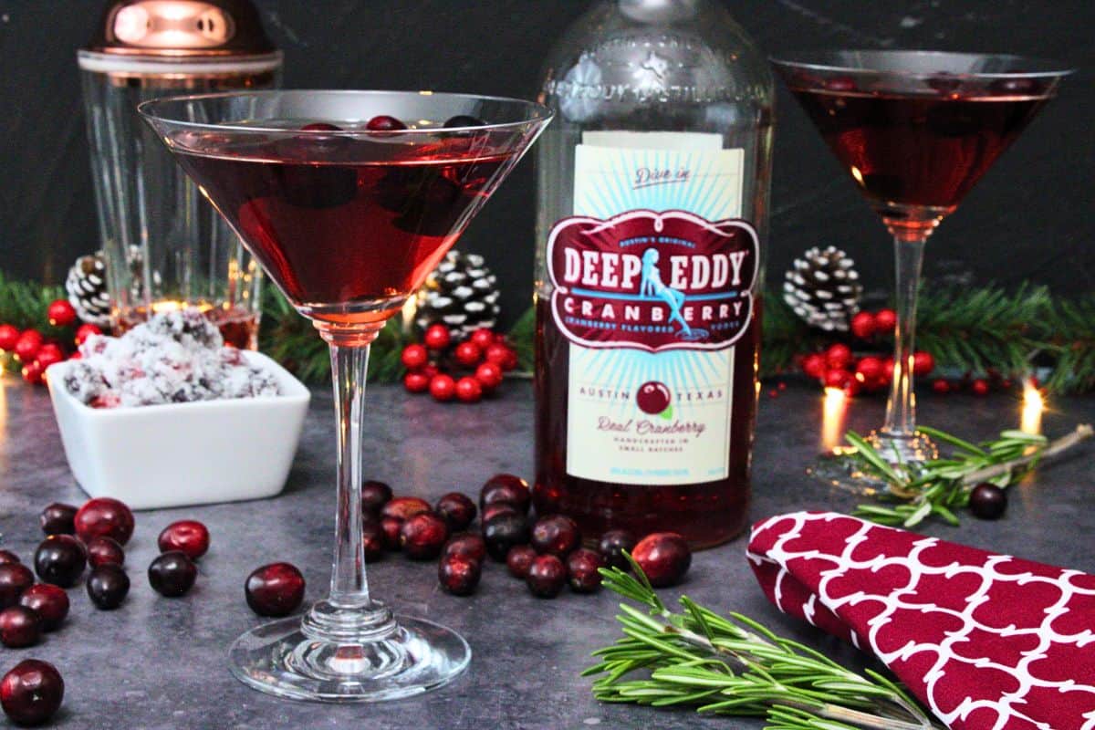 two cranberry martinis with vodka bottle in the center surrounded by fresh cranberries and pine sprigs