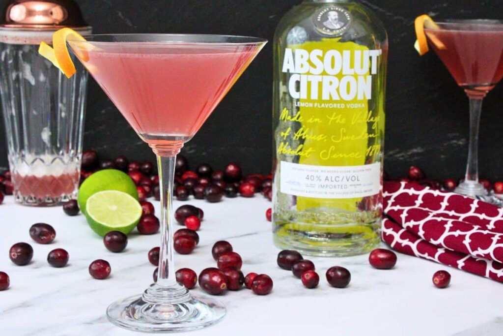 cosmopolitan cocktail in a martini glass with orange rind twist next to vodka bottle and fresh cranberries
