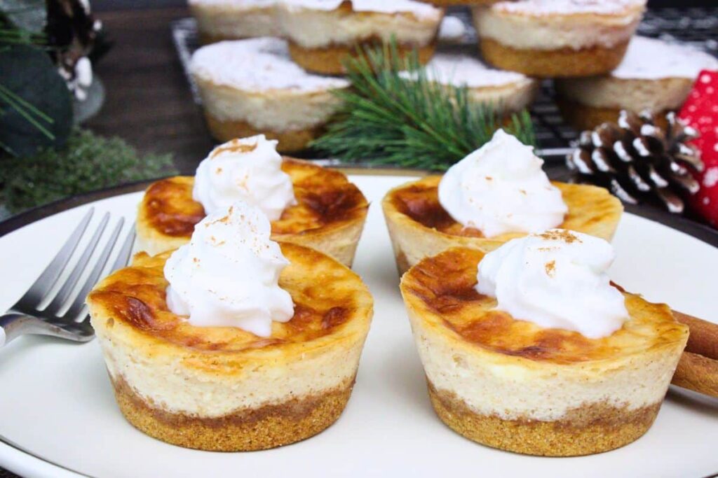four mini eggnog cheesecakes with a swirl of whipped cream on a plate next to a for and winter decorations