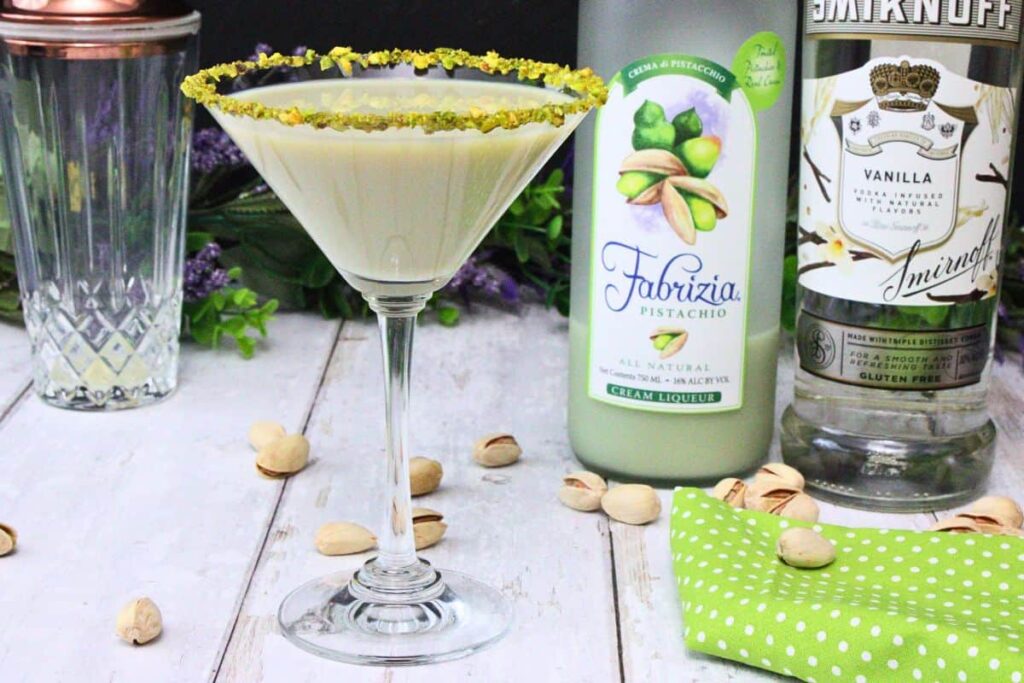 pistachio martini with rimmed glass in front of liquor bottles on a white wooden counter sprinkled with pistachios
