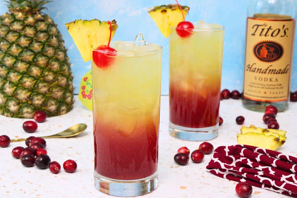 two bay breeze cocktails garnished with pineapple slice and cherry surrounded by fresh cranberries and pineapple