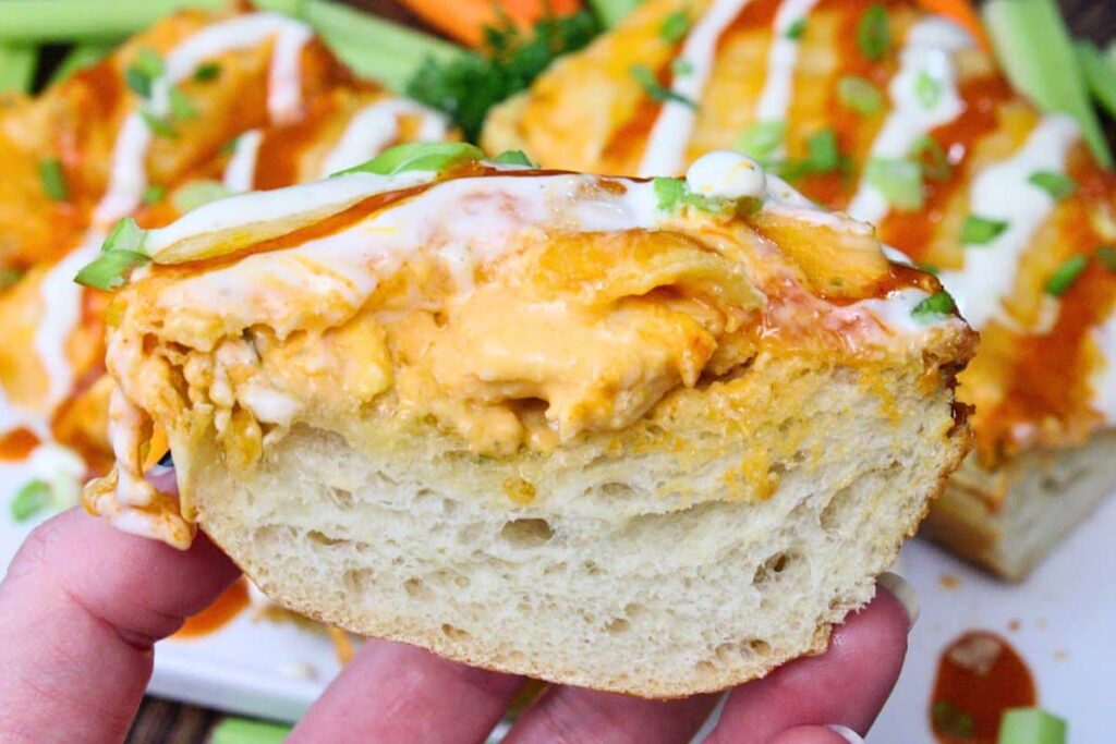 holding up a slice of buffalo chicken garlic bread drizzled with ranch and buffalo sauce showing cheese and shredded chicken