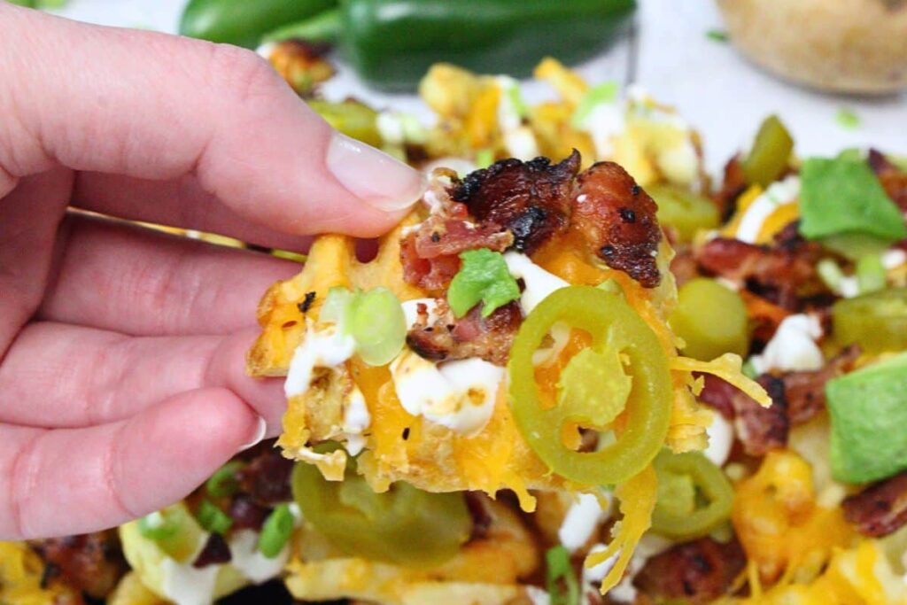 holding up a waffle nacho fry topped with melted cheese, bacon, sour cream, avocado, and jalapeno
