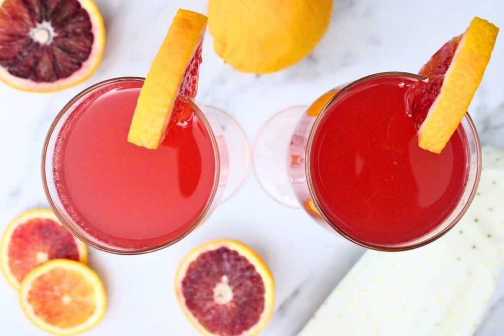overhead view of two blood orange french 75 drinks with blood orange garnish and more fresh fruit surrounding