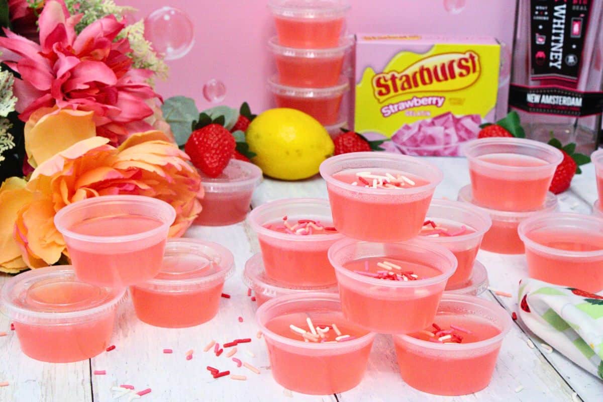 pink whitney jello shots spread and stacked across a white wooden table with flowers, strawberries, and lemons