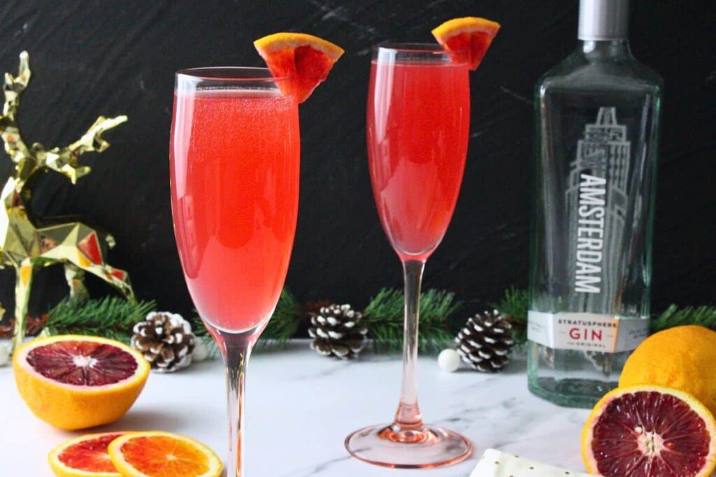two blood orange french 75 cocktails surrounded by fresh fruit in front of gin bottle
