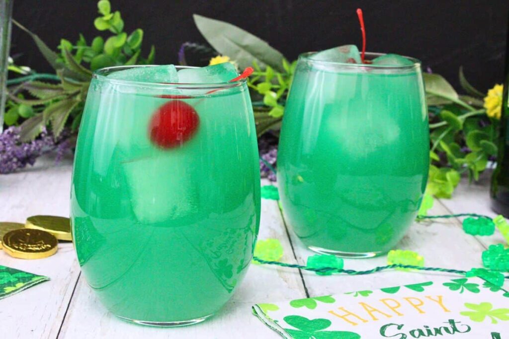 two luck of the irish cocktail garnished with cherry on white wooden table surrounded by st patricks day decorations