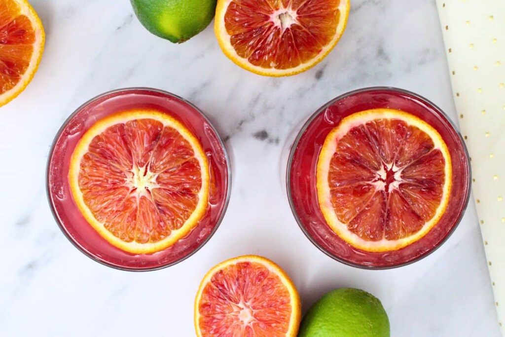 overhead view of two blood orange paloma drinks with blood orange slice floating on top surrounded by fresh fruit