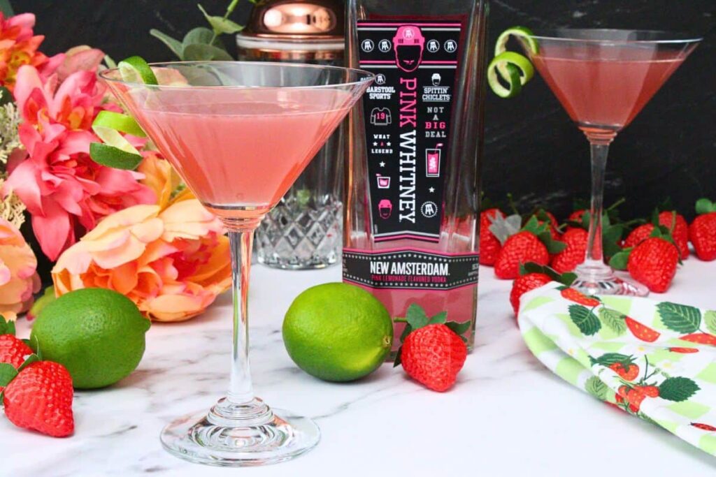 pink whitney cosmo in a martini glass garnished with lime peel in front of vodka bottle