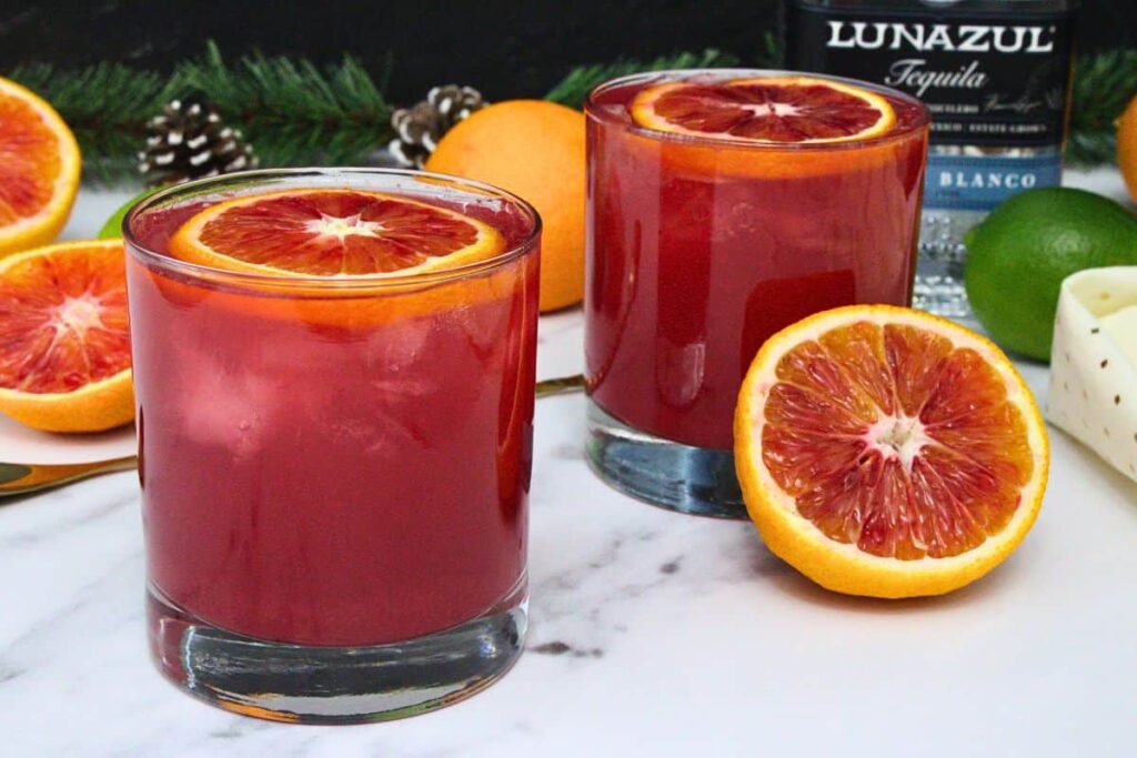 two blood orange paloma cocktails with fresh cut blood oranges surrounding