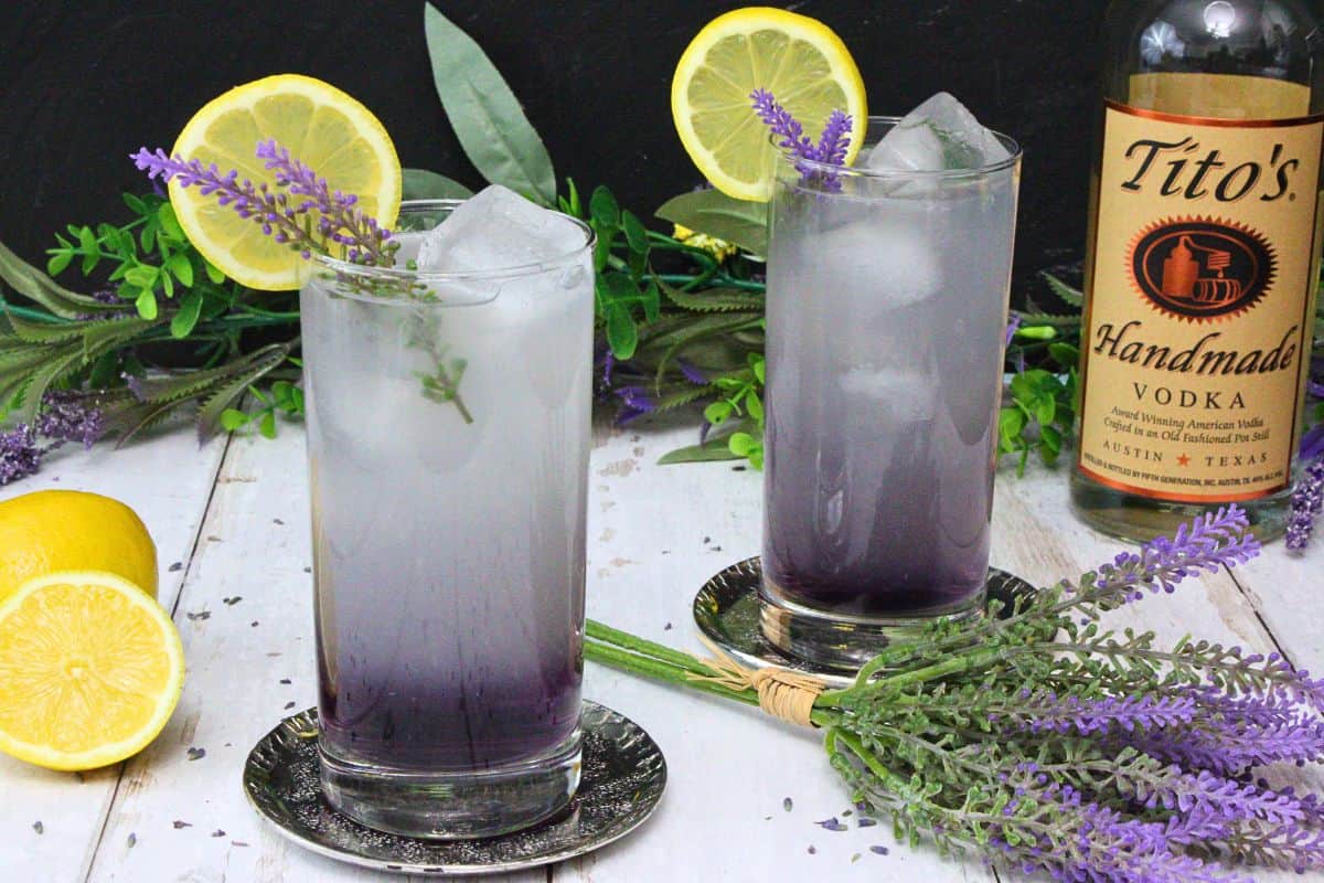 two lavender vodka lemonade cocktails garnished with lavender sprig and lemon slice surrounded by flowers