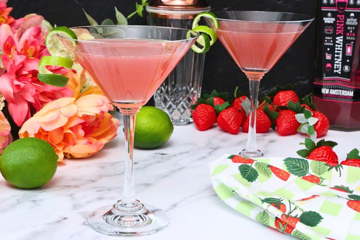 two pink whitney cosmo cocktails with lime peel garnish in martini glasses amongst fruit and flowers