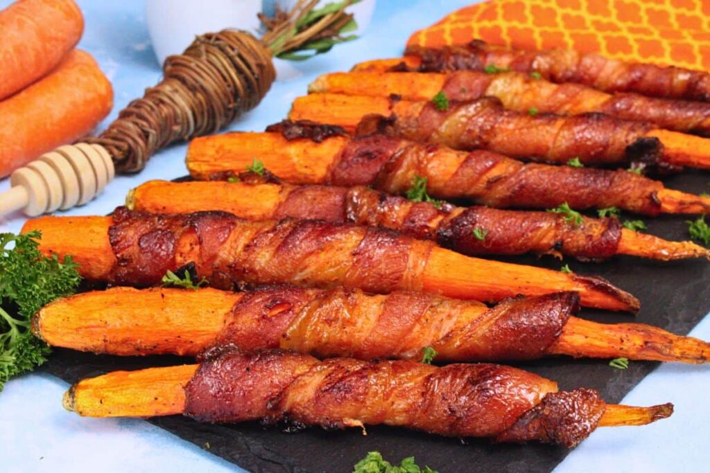 bacon wrapped carrots lined across black slate with fresh carrots in the background
