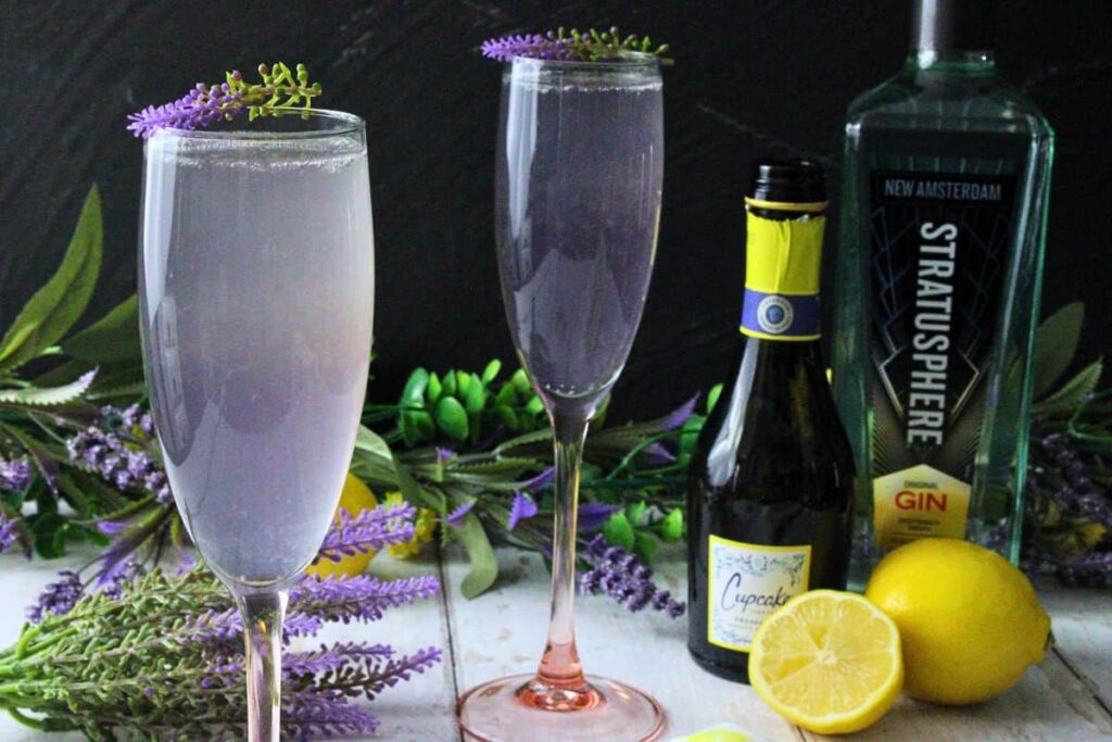 closeup of lavender french 75 garnished with lavender sprig next to a second drink with fresh lemon and bottles
