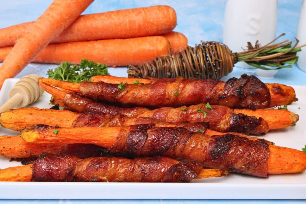 closeup view of bacon wrapped carrots on a white serving plate with fresh carrots in the back