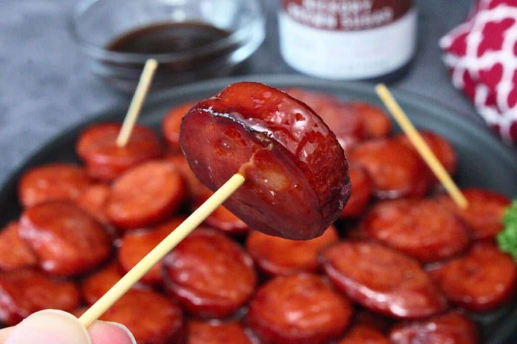 holding up a glazed kielbasa bite on a toothpick over a plate of more bites