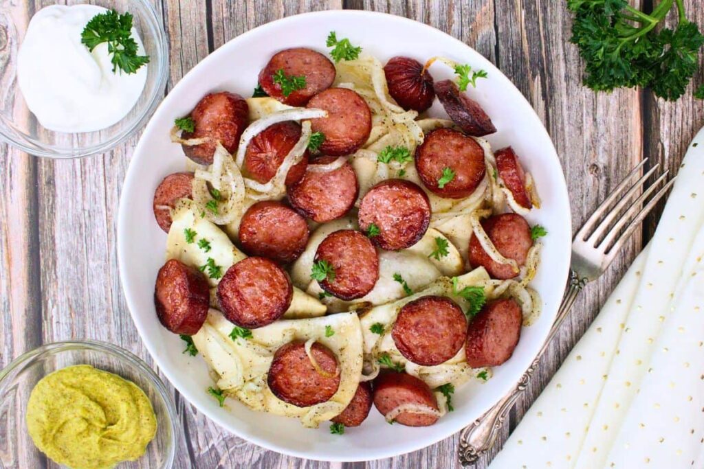 overhead view of kielbasa and pierogies in a bowl with onions and parsley on a wooden table with side of sour cream and mustard