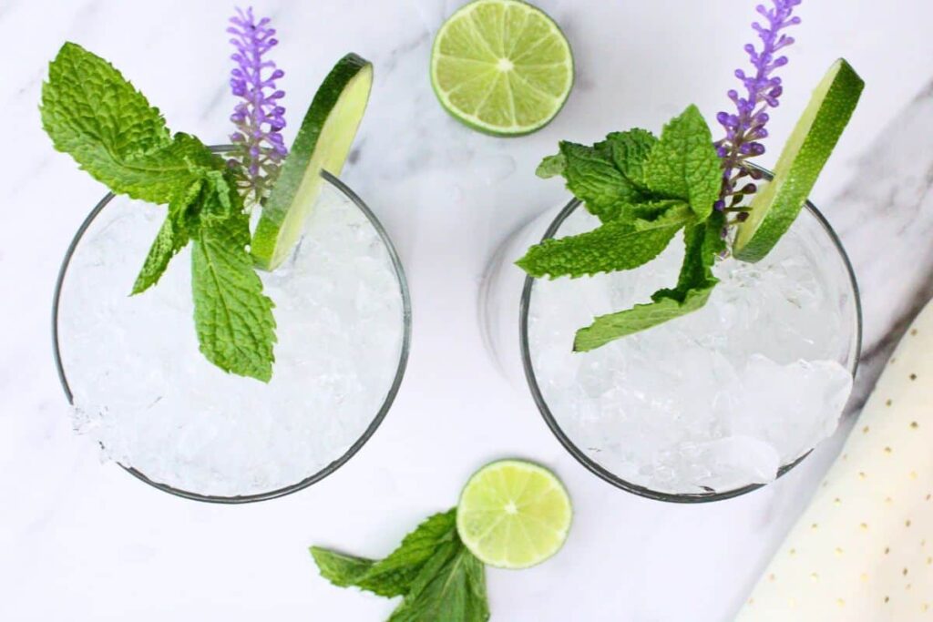 overhead view of two lavender mojito drinks with crushed ice lime and mint on white marble counter