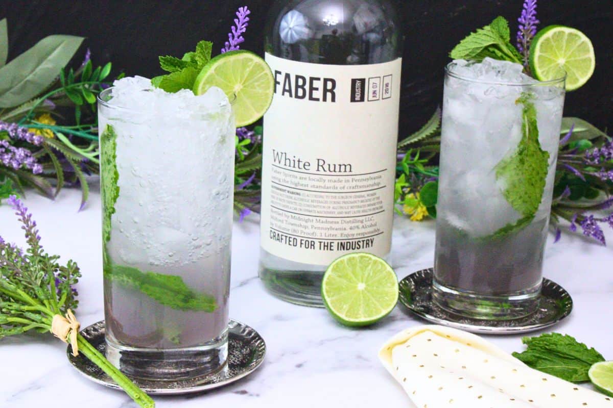 two lavender mojitos in tall glasses with crushed ice garnished with lime wheel and mint surrounding rum bottle