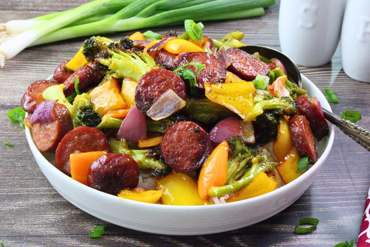 bowl filled with kielbasa stir fry and a serving spoon showing sausage, peppers, broccoli, onions, and rice