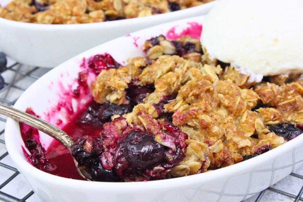 closeup view of blueberry crisp in a dish with a spoonful of the dessert and ice cream scoop on top