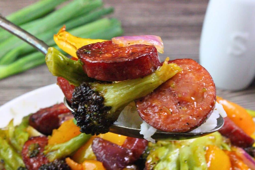 holding up a spoon of kielbasa stir fry showing sausage, broccoli, peppers, onions, and rice