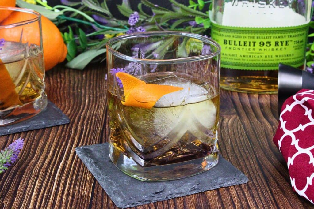 lavender old fashioned with large ice cube, lavender, and orange peel sitting on a slate coaster on a wooden table