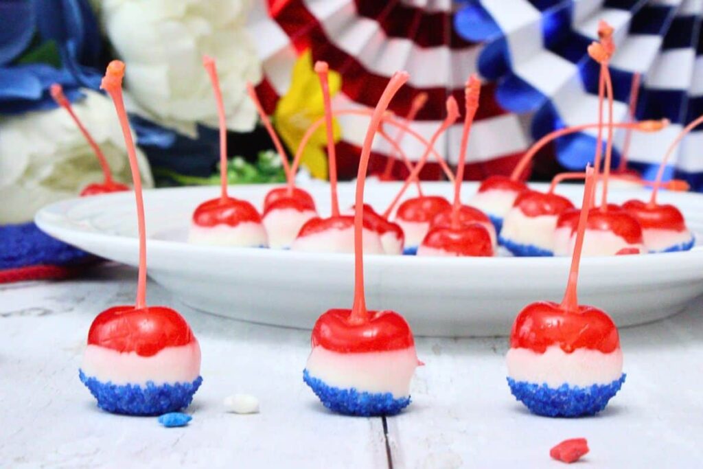 three boozy cherry bombs with red white and blue layers in front of a plate of more cherries and decorations