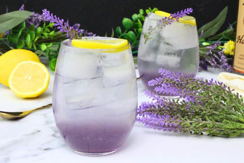 two lavender spritz drinks instemless wine glass garnished with lemon wheel and lavender sprig amongst lemon and flowers